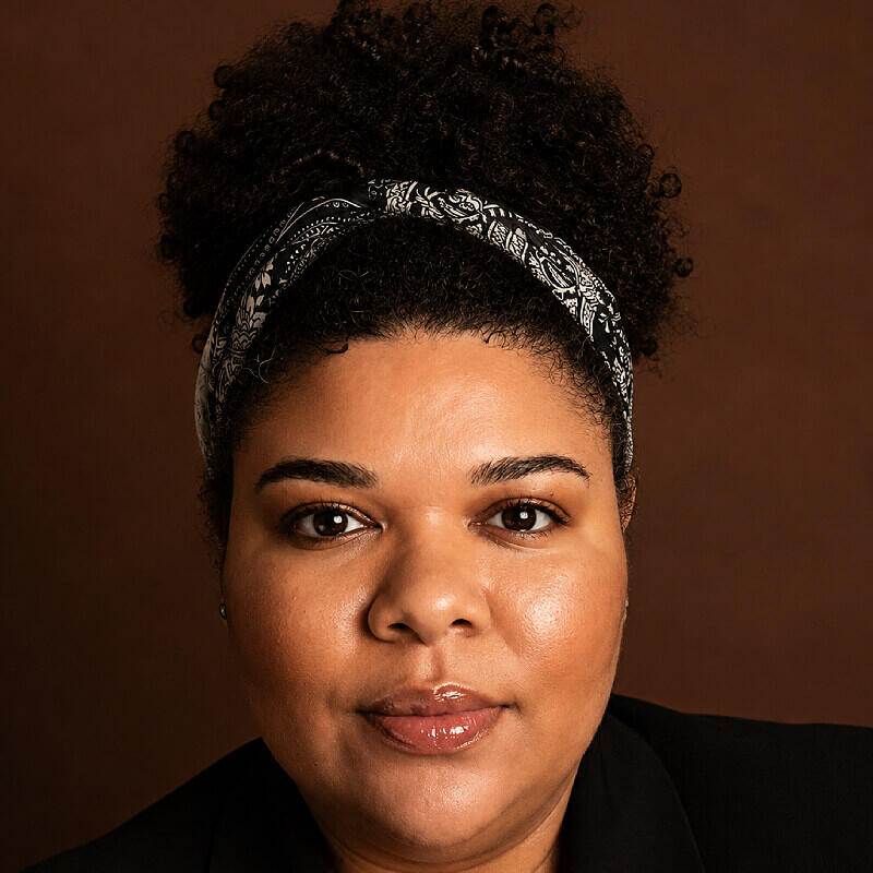 Raquel Neto Retrato em plano fechado de mulher negra com cabelo encaracolado apanhado e fita bandana, com fundo castanho.