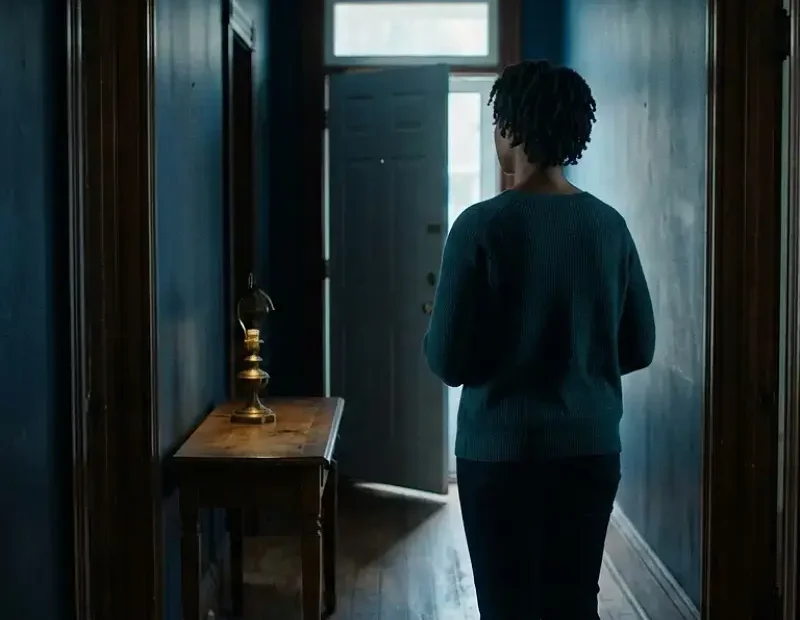 Mulher negra adulta parada num corredor de uma casa pouco iluminada, vista de costas, a olhar para uma porta entreaberta ao fundo por onde entra luz natural, sugerindo contraste entre o interior pesado e a possibilidade de sair para um espaço mais seguro.