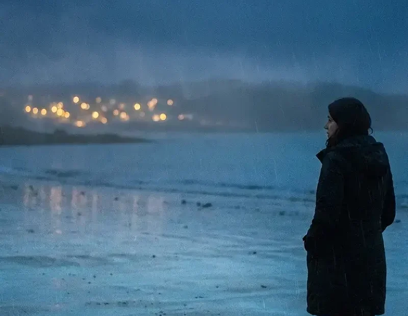 Mulher sozinha à beira-mar numa noite chuvosa, com o mar escuro atrás e reflexos azulados na areia molhada. Ao longe, uma luz dourada desfocada sugere uma festa distante. A imagem transmite solidão, dignidade e a sensação de não ser vista apesar da proximidade dos outros.