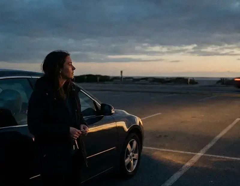 Mulher em pé junto ao seu carro num parque quase vazio ao final da tarde, enquanto outro carro se afasta rapidamente ao longe. A postura é contida e reflexiva, transmitindo abandono, confusão e dignidade após um encontro que terminou de forma abrupta.