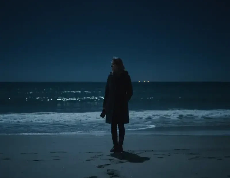 Mulher adulta sozinha numa praia à noite, a segurar um telemóvel com o ecrã apagado, olhando para o mar.