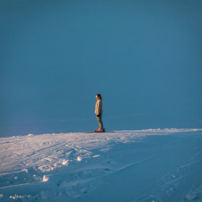 Uma mulher parada no topo de uma pista de ski, sem equipamento adequado, a olhar para baixo com expressão meio incrédula, meio resignada. O cenário é amplo, frio, com céu azul pálido. A imagem sugere a “descida” da meia-idade feita sem manual de instruções.
