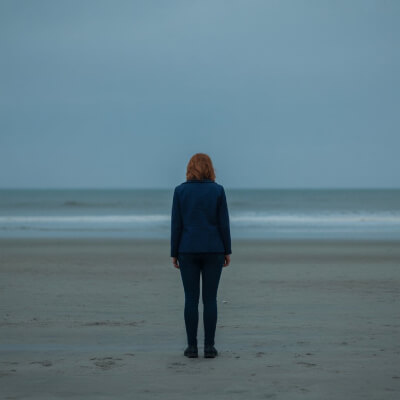 Mulher sozinha numa praia ampla. O céu está nublado. Atmosfera de espera, contenção e desalinhamento interno.