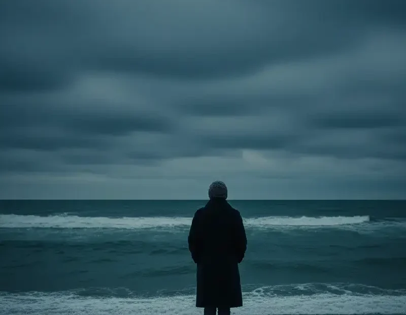 Uma mulher sozinha junto ao mar no inverno, de costas para a câmara. O céu está pesado, nublado, e o mar agitado, com ondas irregulares que não convidam a entrar. A figura transmite suspensão e desalinhamento interior: não avança, não recua. As cores são frias e profundas, com azuis escuros e médios, e um apontamento discreto de dourado na luz difusa, sugerindo resistência mais do que esperança. A imagem evoca perda de rumo, desgaste emocional e a sensação de estar parada enquanto o tempo continua a passar.